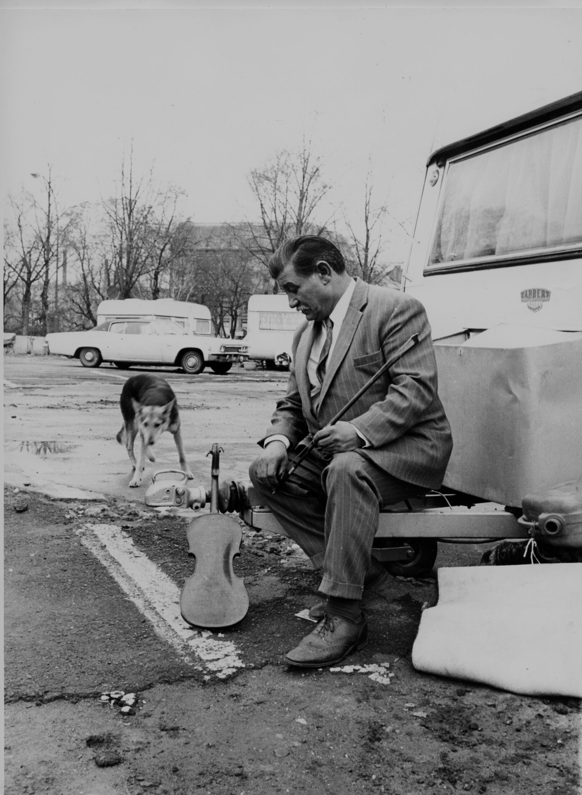 En mann utenfor en campingvogn p&aring; asfalt, han holder en fiolin, det er en hund i bakgrunnen.